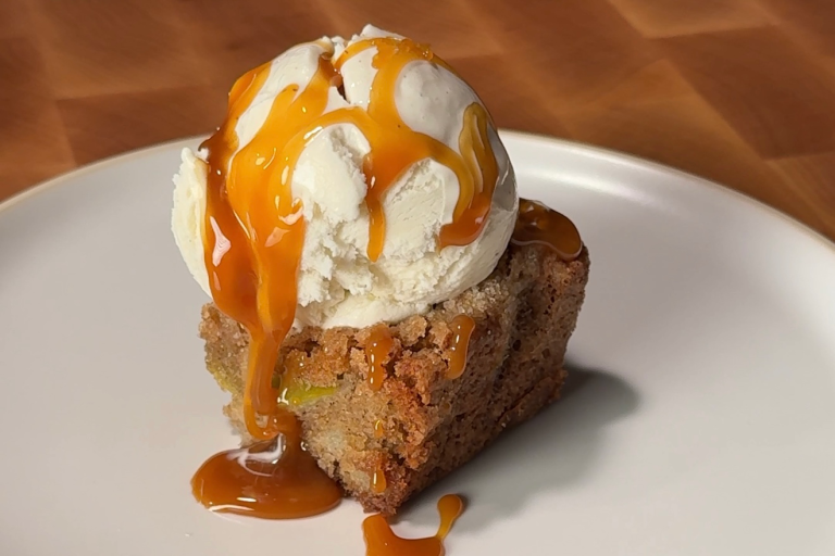 fresh apple cookie bar topped with ice cream and drizzled with caramel on a white ceramic plate over a wooden cutting board