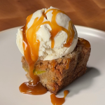 fresh apple cookie bar topped with ice cream and drizzled with caramel on a white ceramic plate over a wooden cutting board with candled background